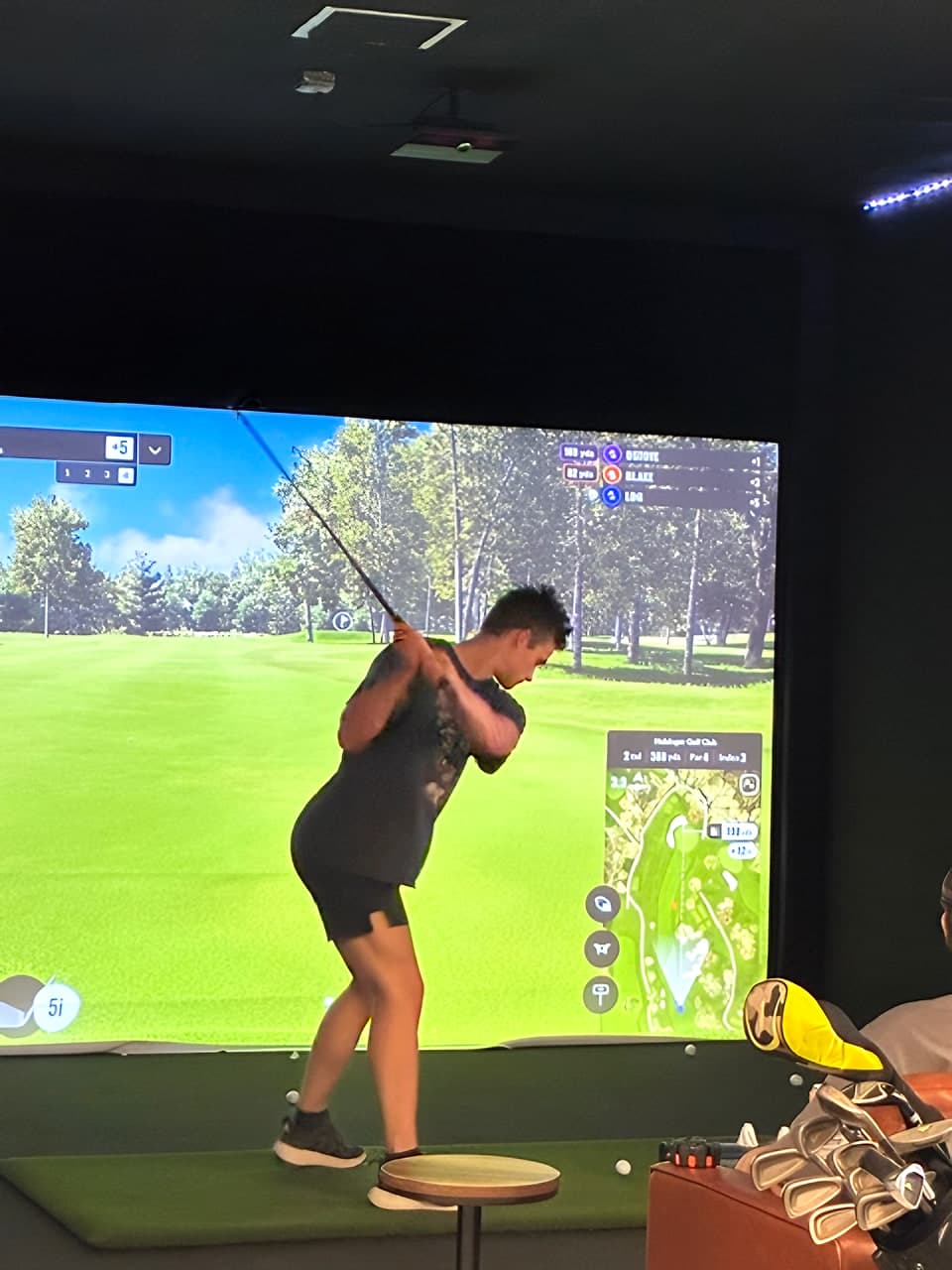 A golfer follows through on a full swing in front of a Trackman screen showing a tree-lined fairway