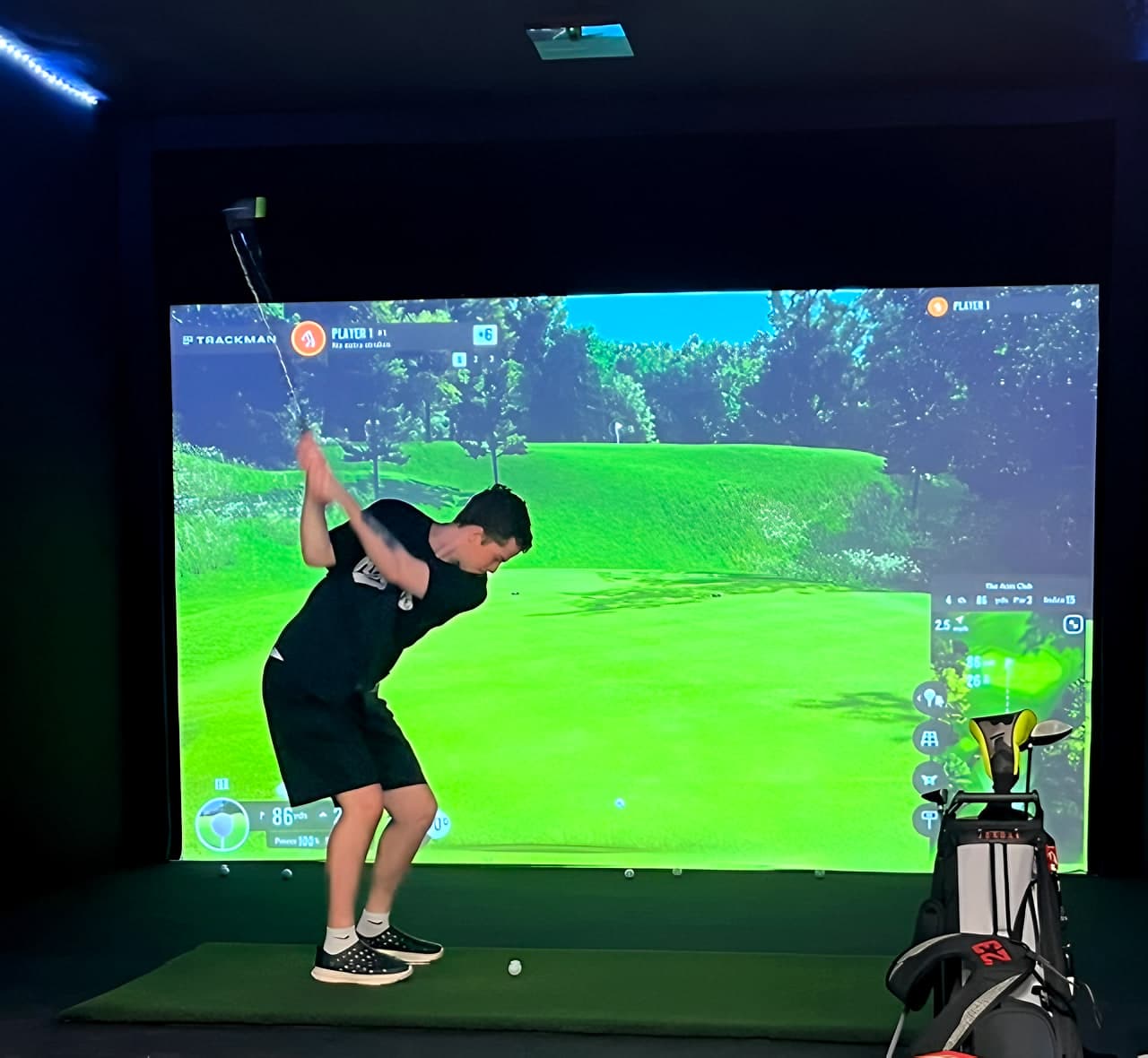 A golfer at the top of the backswing on the hitting mat, a Trackman round on the screen and a golf bag beside the bay
