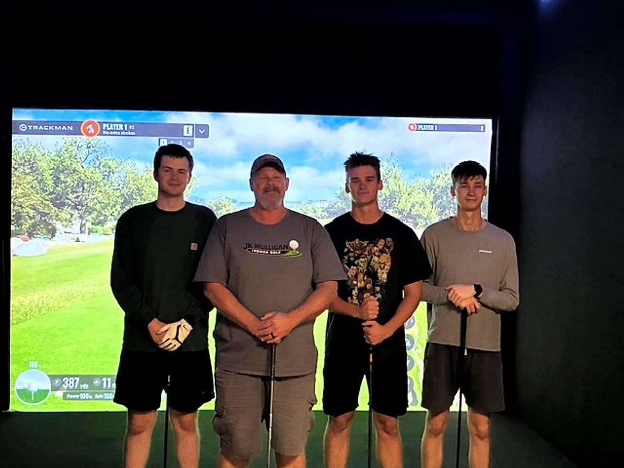 A group of four golfers holding their clubs and posing in front of a Trackman simulator screen