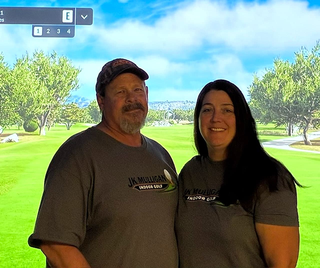 Two people in JK Mulligan Indoor Golf shirts smiling in front of a simulated golf course