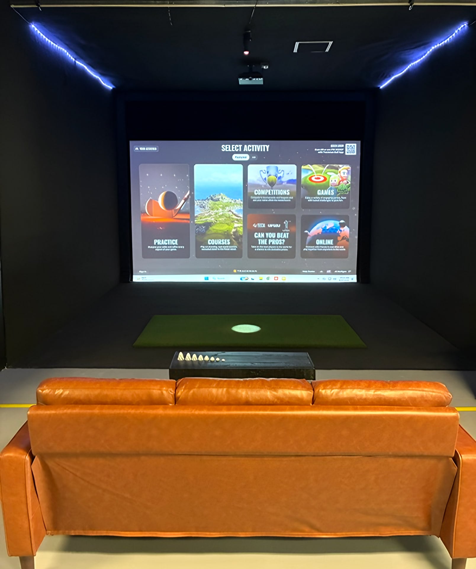 A JK Mulligan simulator bay: the Trackman screen showing its activity menu above the hitting mat, with a leather couch for your group in the foreground